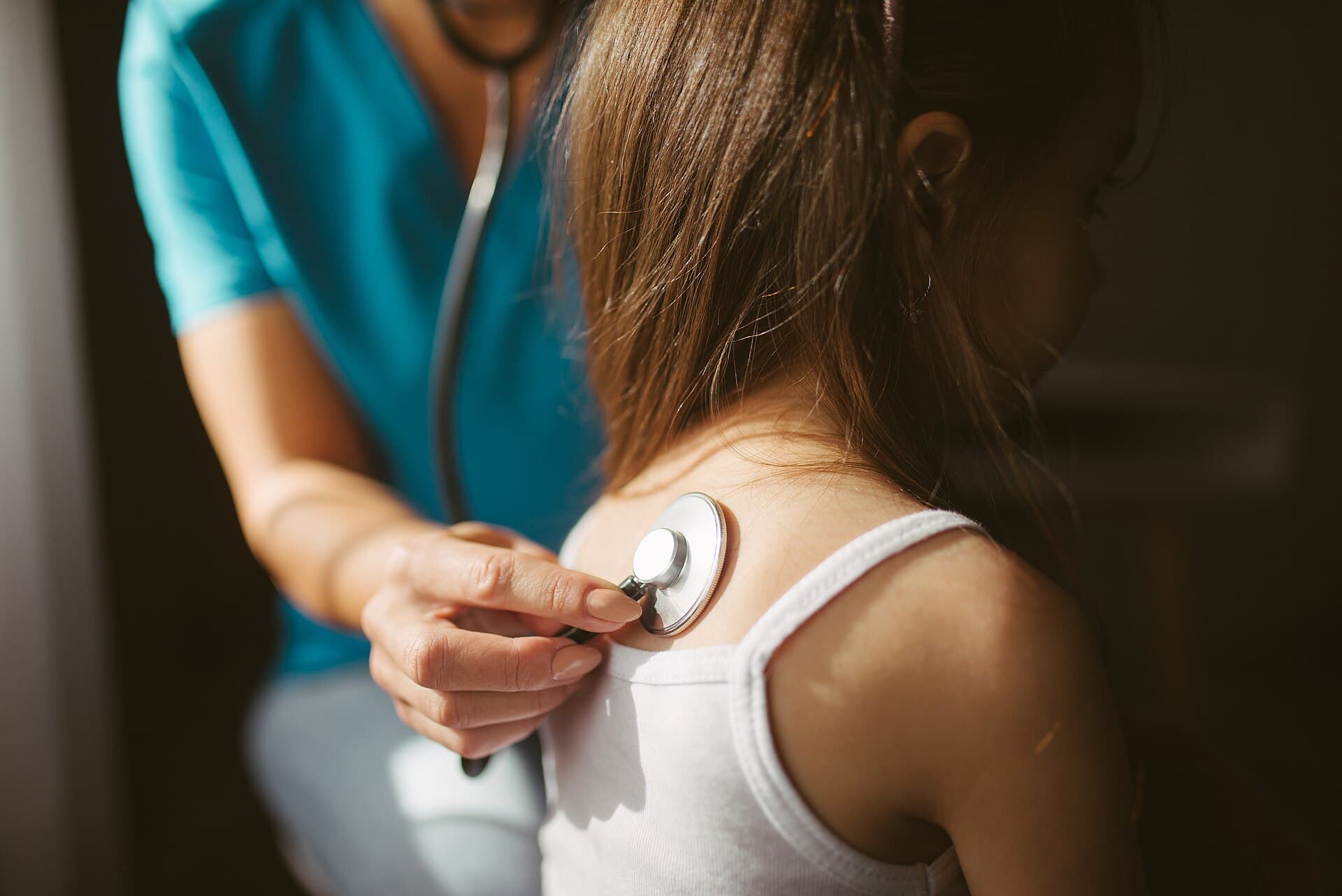 Médica de uniforme azul encosta o estetoscópio nas costas de uma paciente, em close.
