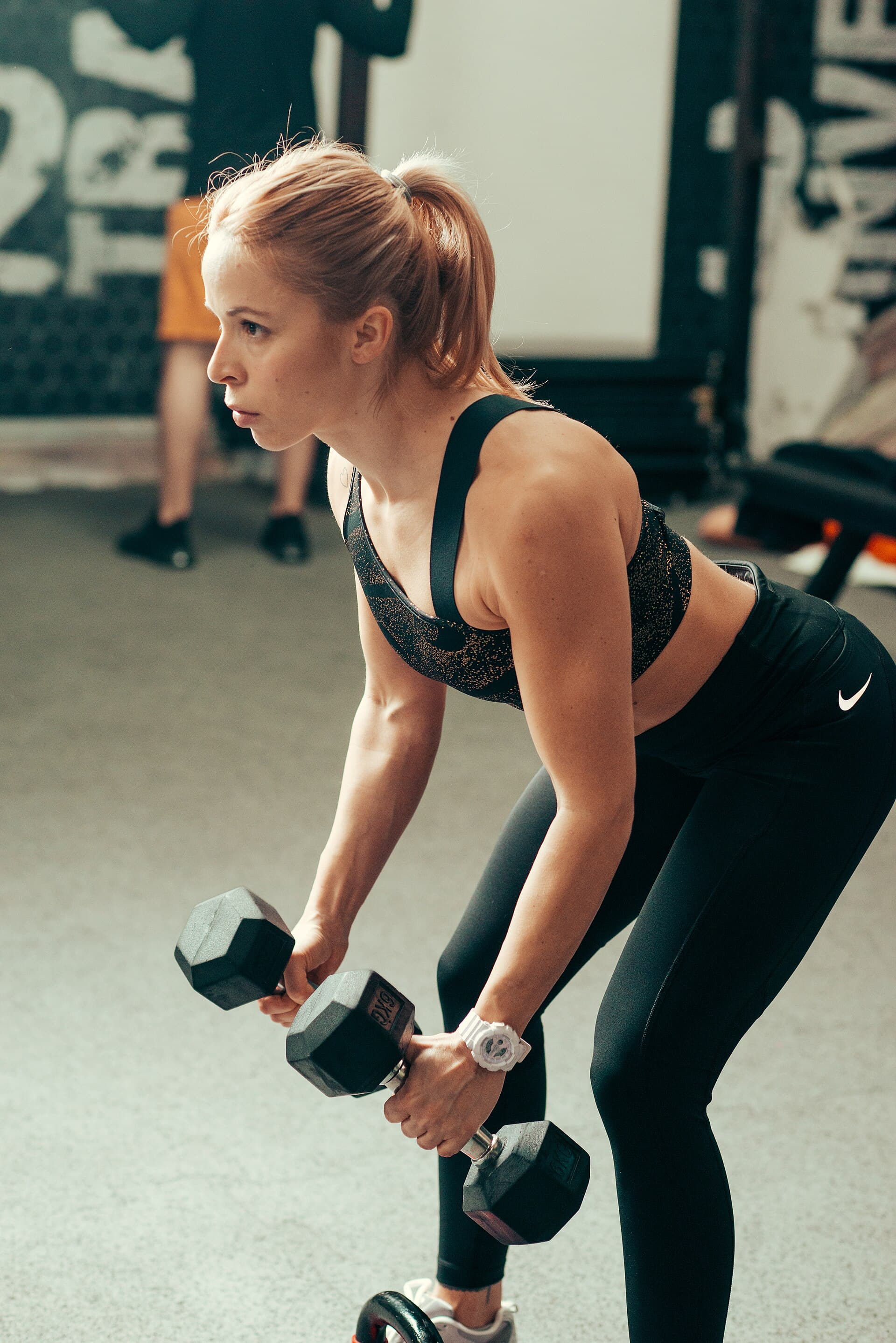 Mulher de roupa de treino escura levanta halteres em uma academia, concentrada no movimento.