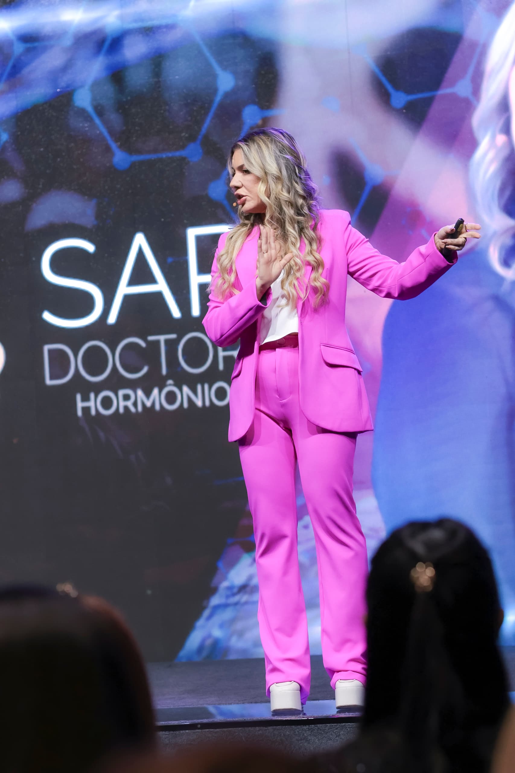 Dra. Sarina Occhipinti no palco de um evento da SariDoctors, de terno rosa, falando ao público com um telão ao fundo.